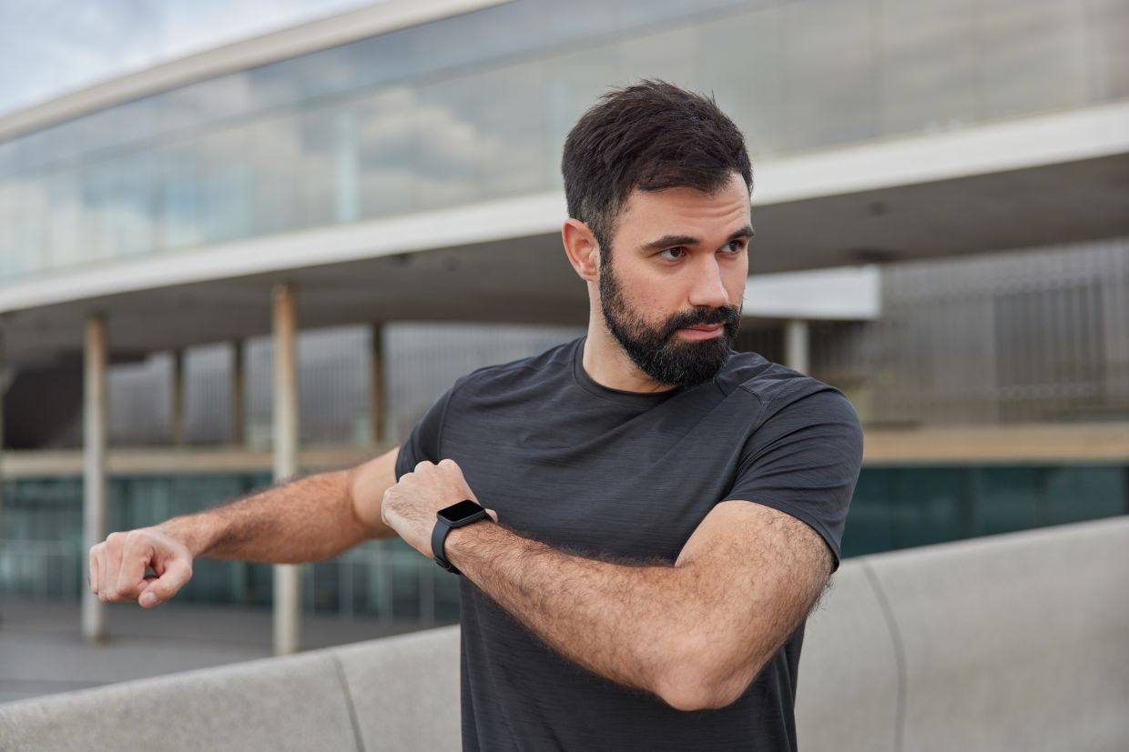 Outdoor shot of motivated bearded man stretches arms warms up before workout wears smartwacth casual black t shirt concentrated into distance poses against blurred background. Sport concept.