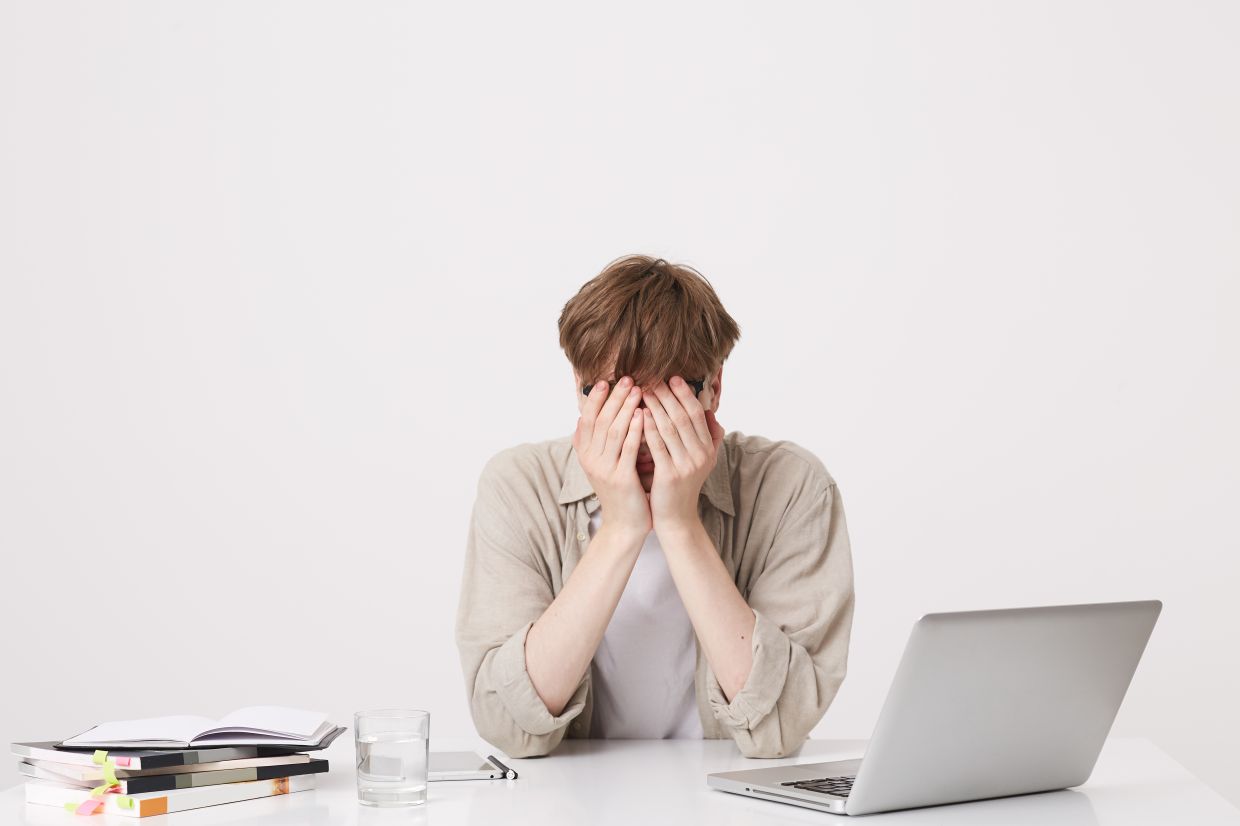 Closeup of cheerful young male student with braces wears beige shirt study using laptop computer and notebooks sitting at the table isolated over white background