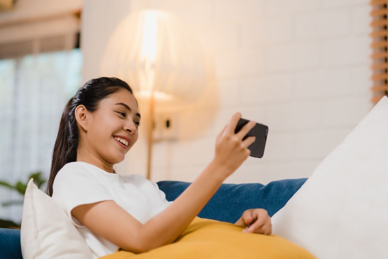 Young Asian woman using smartphone checking social media feeling happy smiling while lying on the sofa when relax in living room at home. Lifestyle latin and hispanic ethnicity women at house concept.