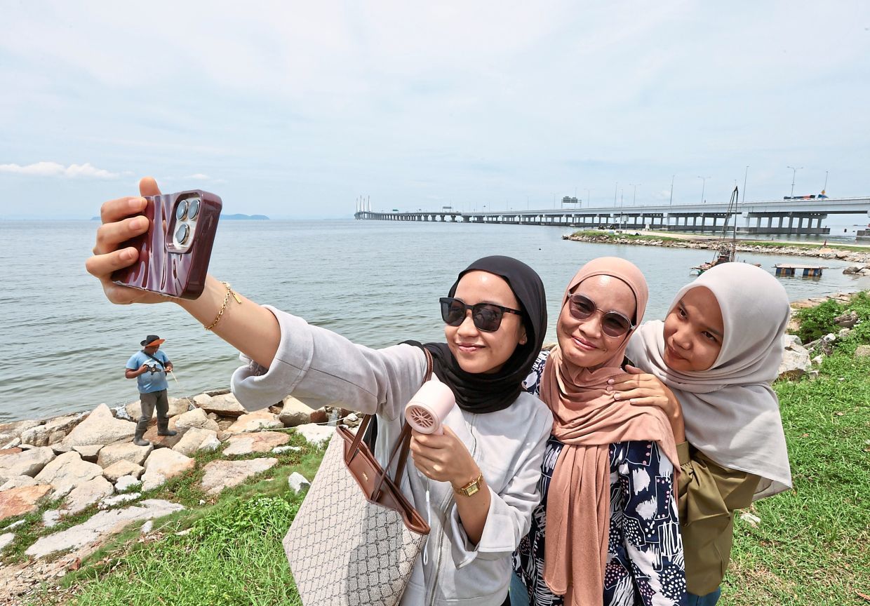 Halimah Munthe,23 wearing sunglasses and using portable fan takes selfies with her mother and sister near Penang Bridge as the sun peaks in Bayan Lepas in Penang. (July 22, 2025) — ZHAFARAN NASIB/The Star.