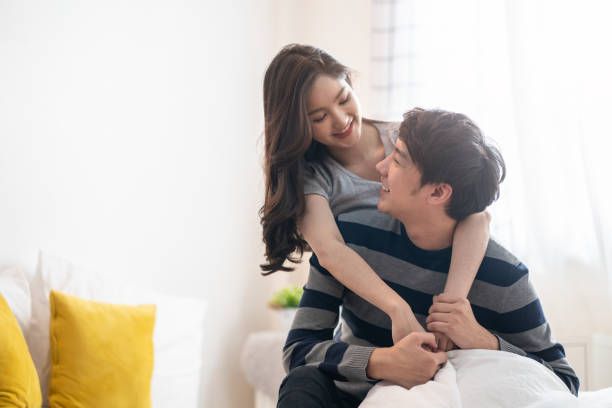 Asian young couples man and woman staying together in bed room. Girl hugging boy from behind, looking eyes at each other feeling love happy with smile face. Relationship of young girl and boy concept