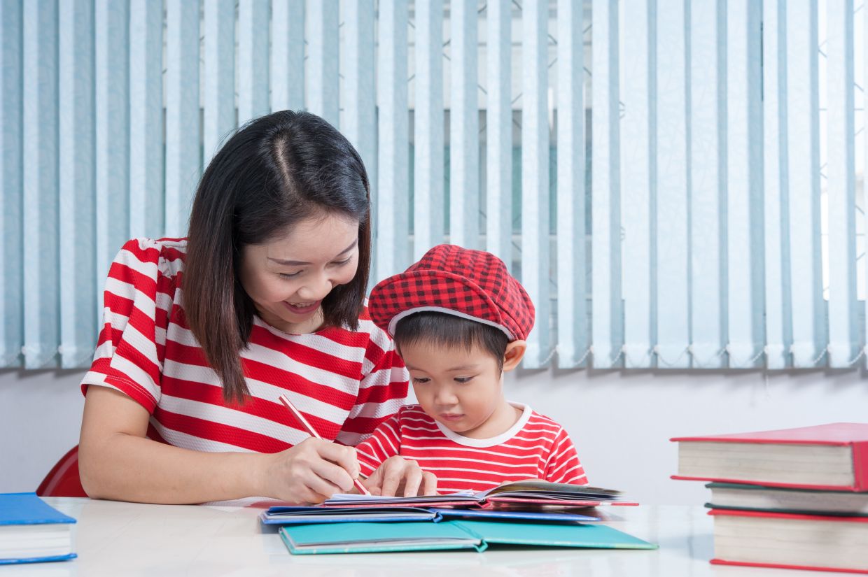 Asian cute boy doing his school homework with his mother, at home, he is writing on a book