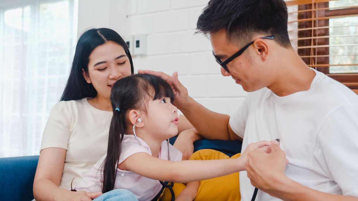 Happy cheerful Asian family dad, mom and daughter playing funny game as doctor having fun on sofa at home. Self-isolation, stay at home, social distancing, quarantine for coronavirus prevention.