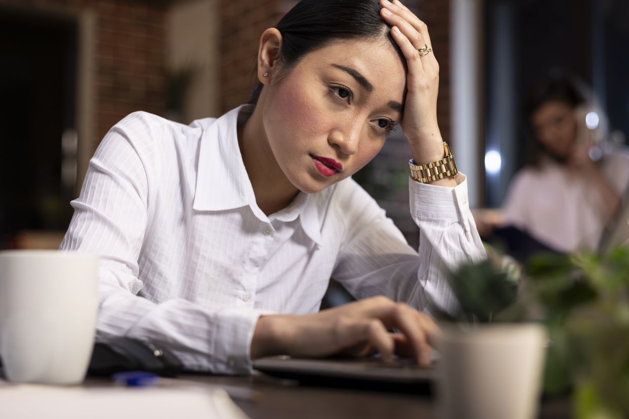 Fatigued asian manager holding her forehead, overwhelmed by deadlines, browsing on her laptop late at night. Closeup of woman looking exhausted as she sits at desk, trying to complete business tasks.
