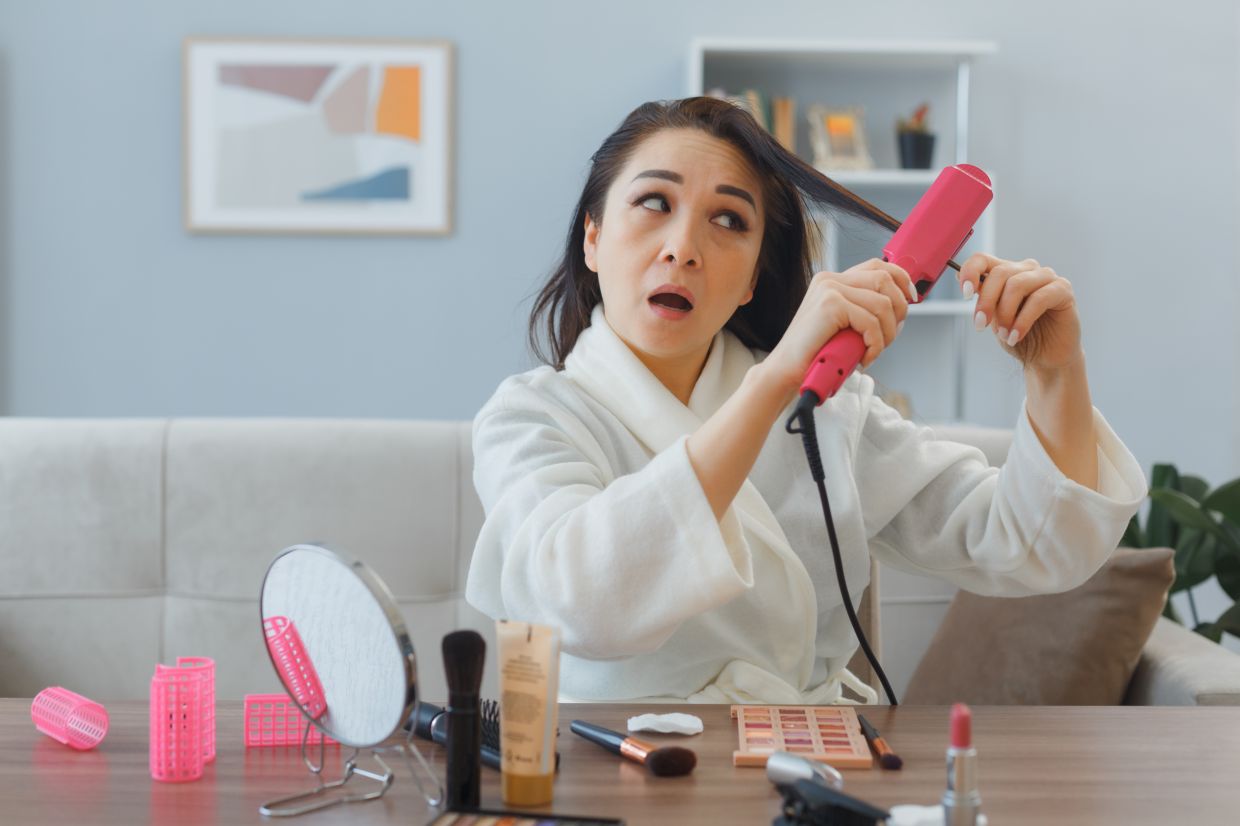 young asian woman with towel in bathrobe sitting at the dressing table at home interior using curling ironmaking styling being confused doing morning makeup routine