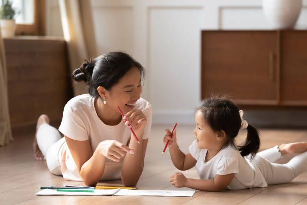 Educational pastime develop creativity skill in kid concept. Asian mother her small daughter lying on warm wooden floor in sunny cozy living room, mom teach girl paint use album and colourful pencils
