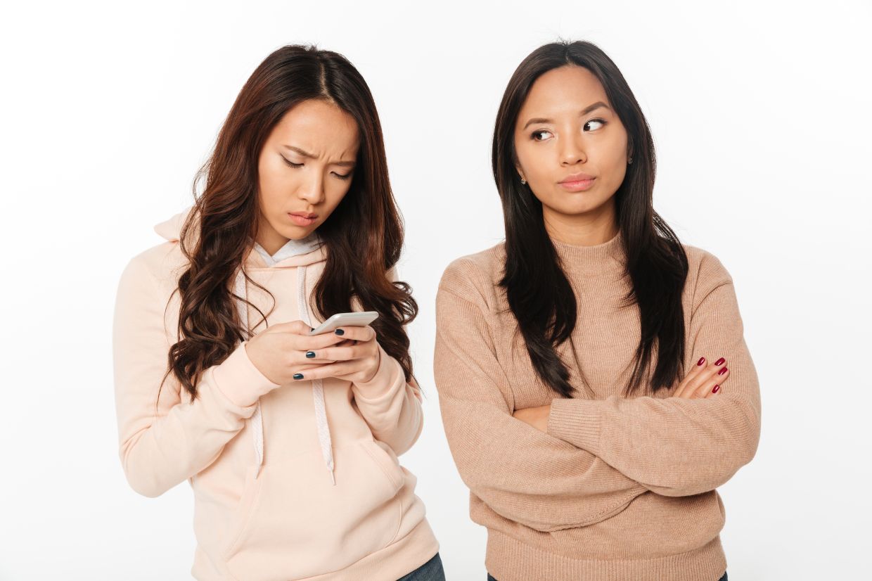 Photo of asian girl displeased because of her sister chatting by mobile phone. Looking aside.