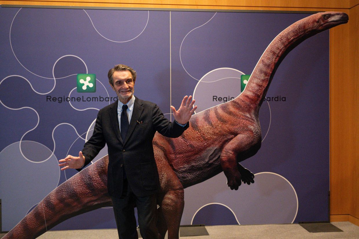 Attilio Fontana, President of Lombardia Region, poses for a photograph with a cardboard dinosaur during a press conference to present the discovery of thousands of dinosaur tracks discovered in Italy's Stelvio National Park near the areas that will host the Milano-Cortina 2026 Olympic Games, in Milan on December 16, 2025. The tracks are estimated to belong to Late Triassic prosauropod dinosaurs, dating back 210 million years. (Photo by Piero CRUCIATTI / AFP)