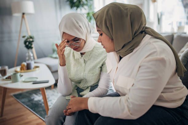 Young Indian woman consoling her sad friend while sitting on sofa together in living room.